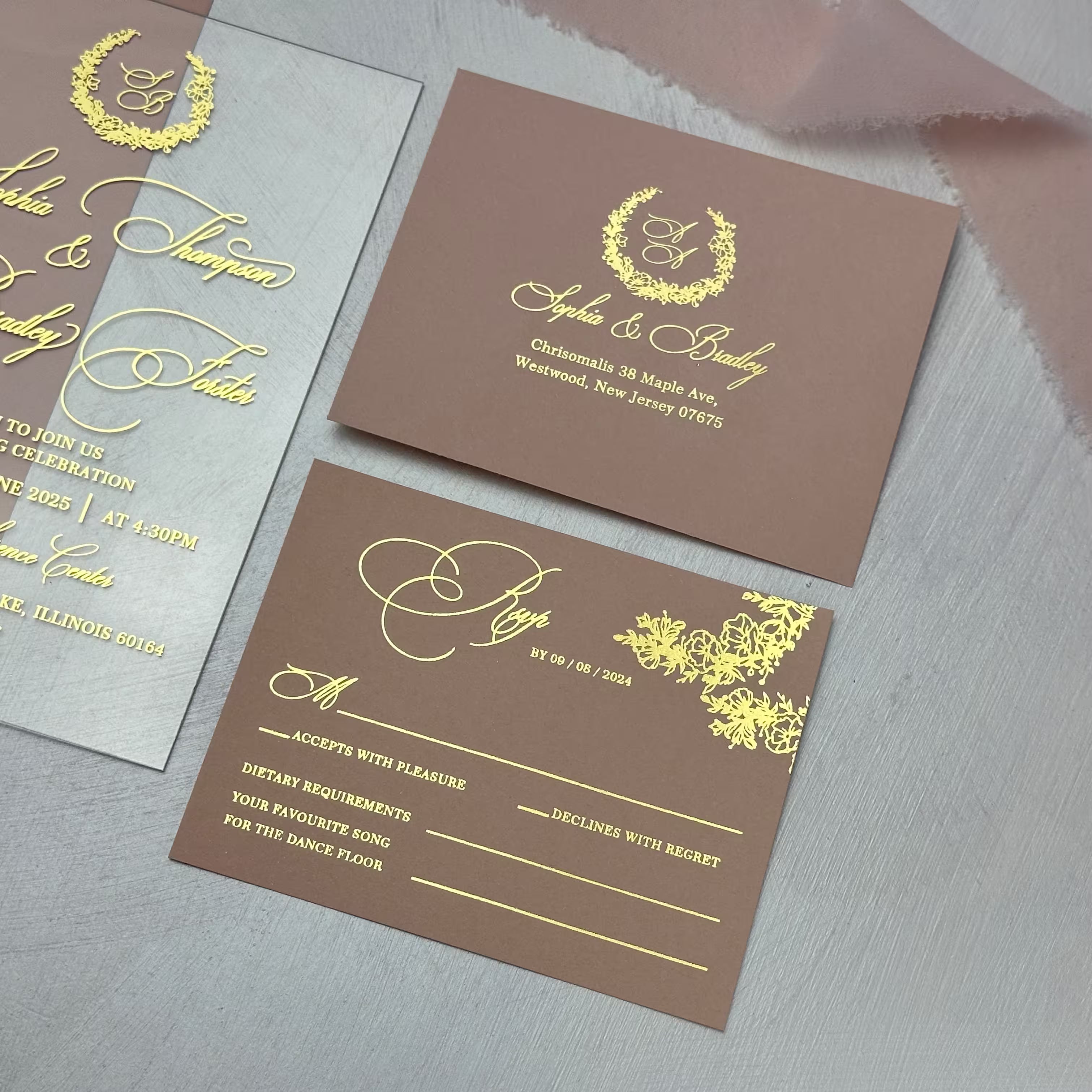 Clear acrylic wedding invitation with gold foil calligraphy beside coordinating RSVP and address cards printed with a monogram wreath
