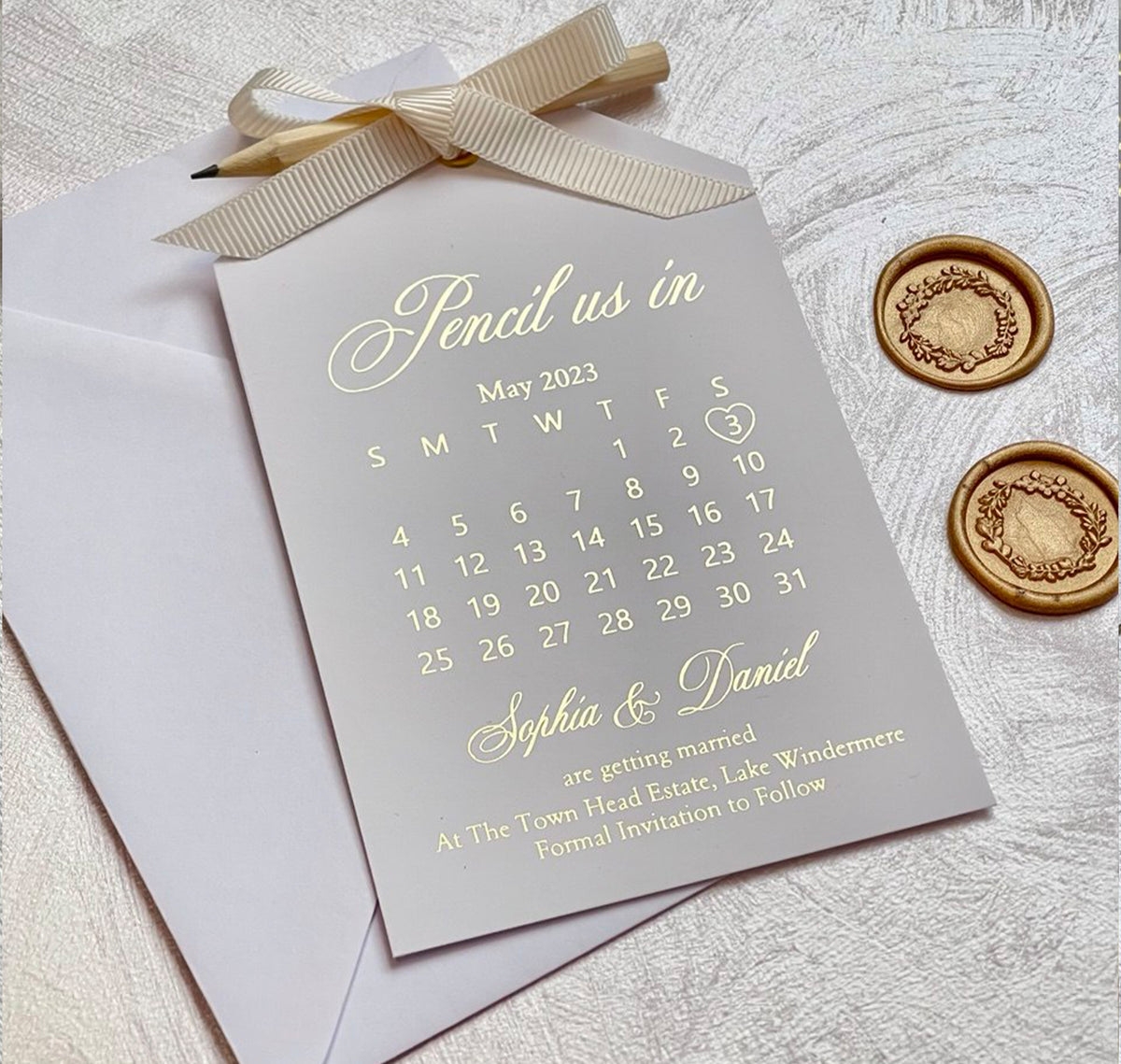 Pencil us in save the date cards showing a month calendar in foil, tied with a ribbon and a pencil, personalized for the couple.