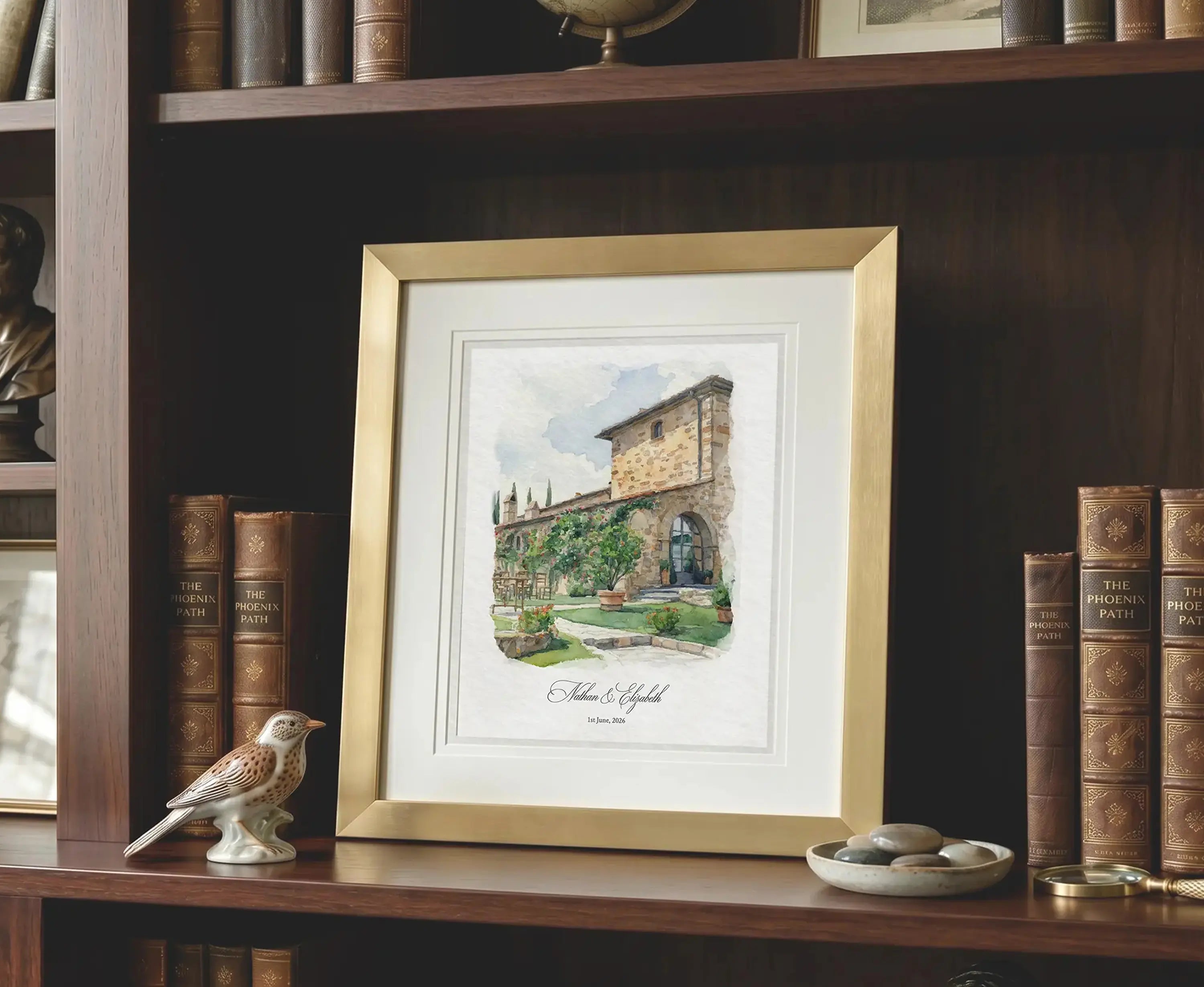    Watercolor venue portrait displayed in a matte gold frame with white matting, styled on a classic wooden bookshelf in a traditional interior setting.