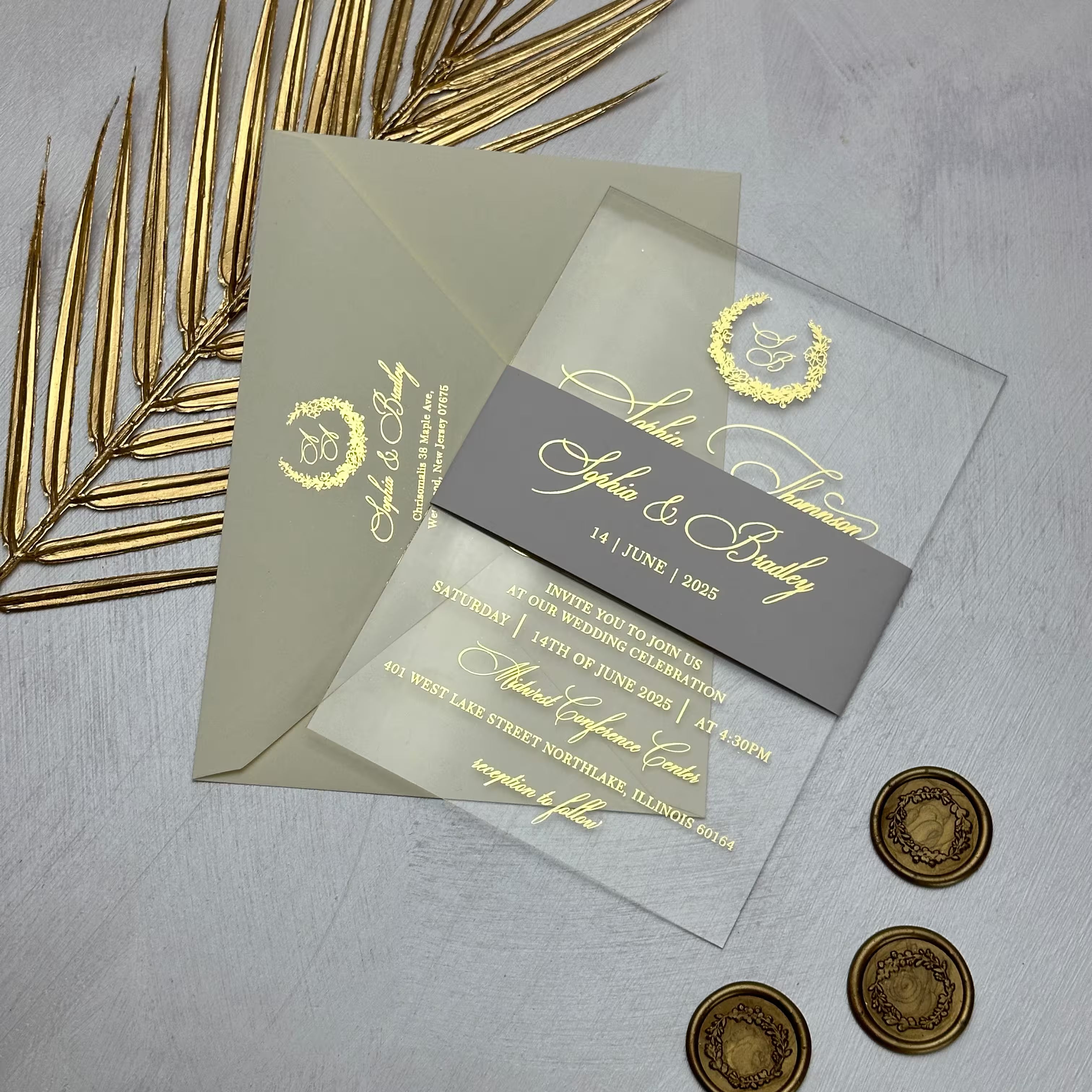 Clear acrylic wedding invitation with a gold foil monogram wreath, held by a grey belly band and set against a printed envelope with the couple's monogram