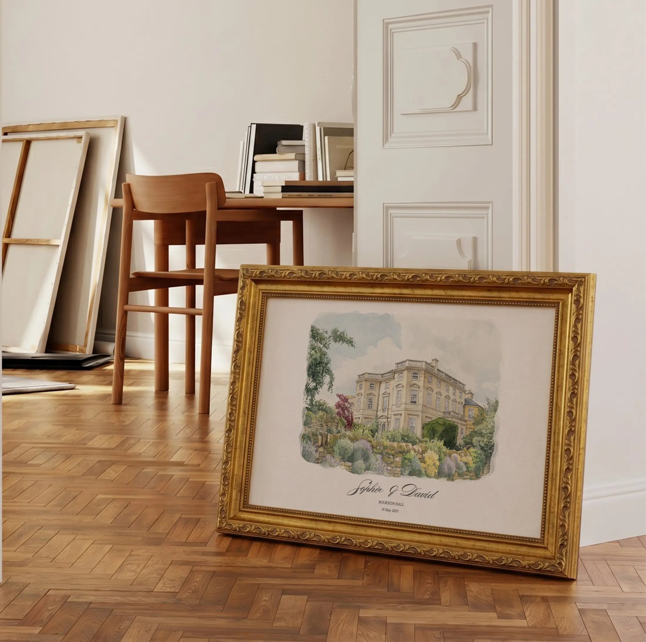 Framed watercolor wedding venue portrait displayed in an elegant interior with warm wooden flooring and soft natural light. The artwork features a hand-painted architectural illustration of a classical manor house, presented in an ornate antique-style gold frame with decorative detailing. Personalized names and wedding date are printed beneath the artwork, creating a refined and timeless presentation suitable for luxury interiors and meaningful wedding keepsakes.