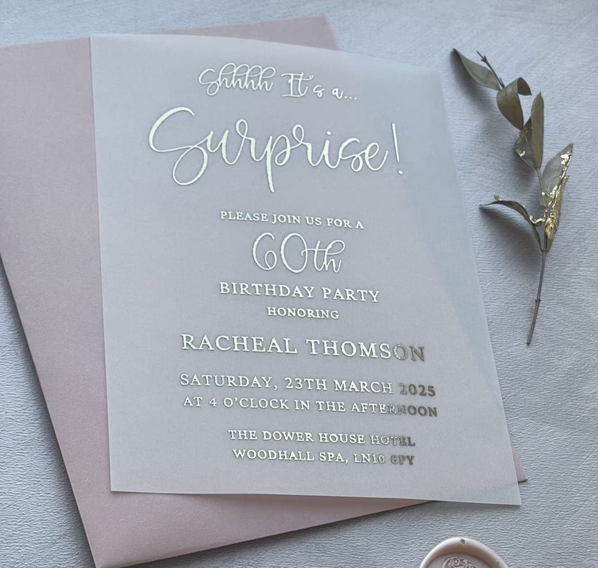 Surprise party invitations on translucent vellum with foil lettering, personalized surprise birthday invitation shown with an envelope and a dried sprig.